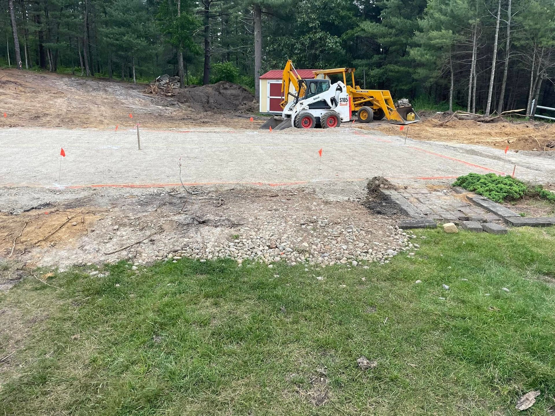 Construction site with gravel road, skid steer and backhoe. Green grass in foreground, trees in background.