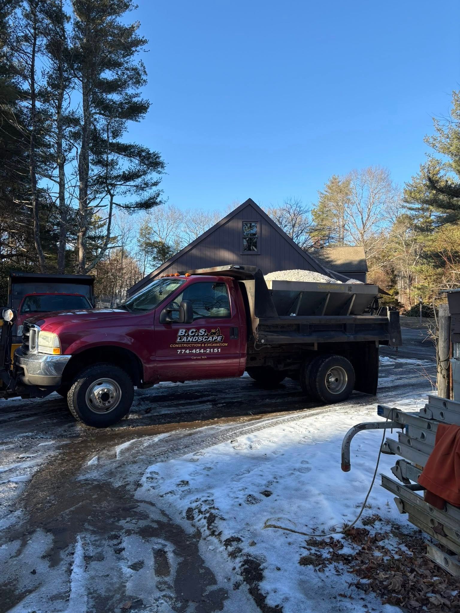 Red dump truck parked on snowy ground, in front of a house. The truck bed is full. Clear, blue sky.