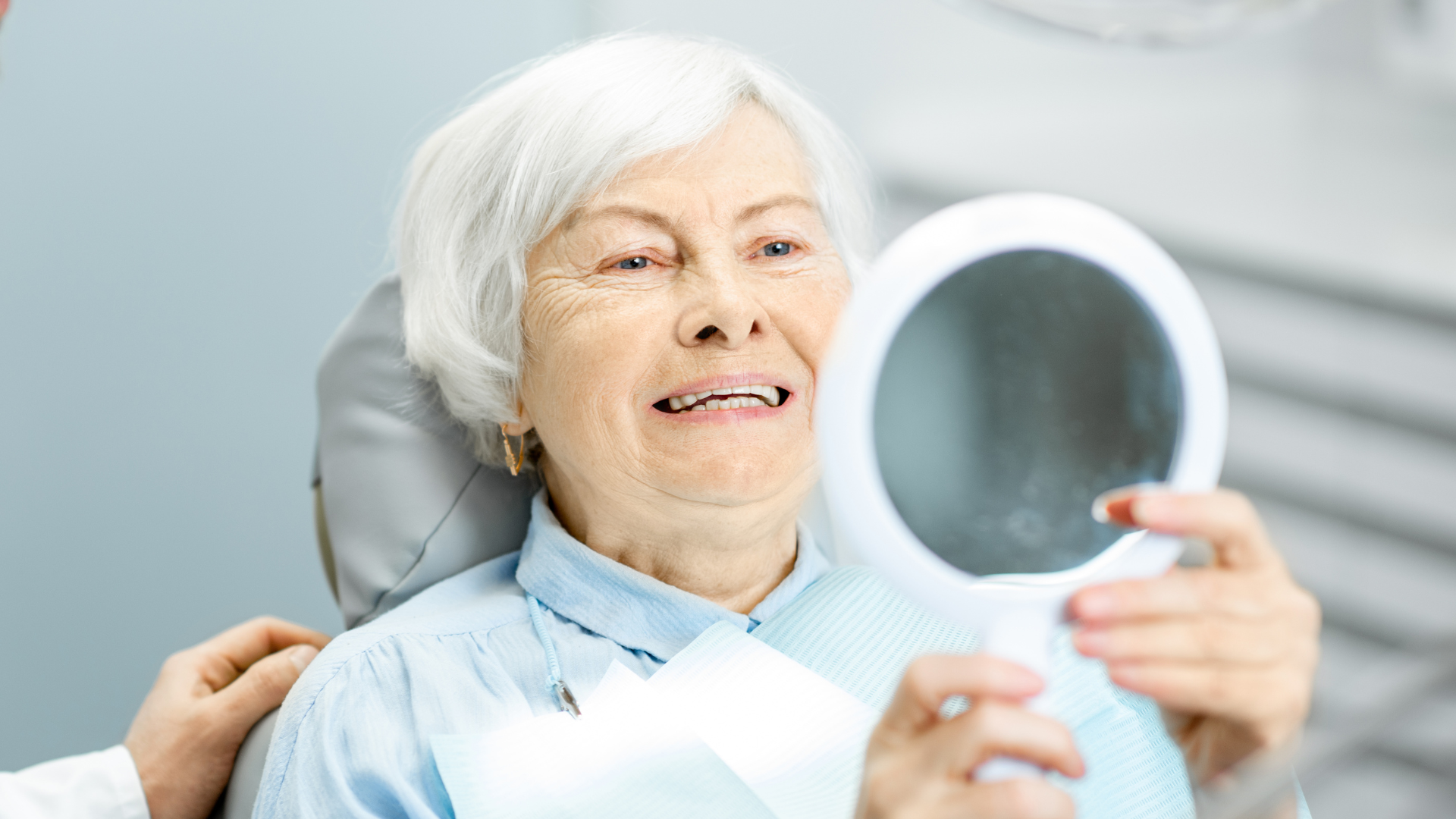 An elderly woman is looking at her teeth in a mirror.