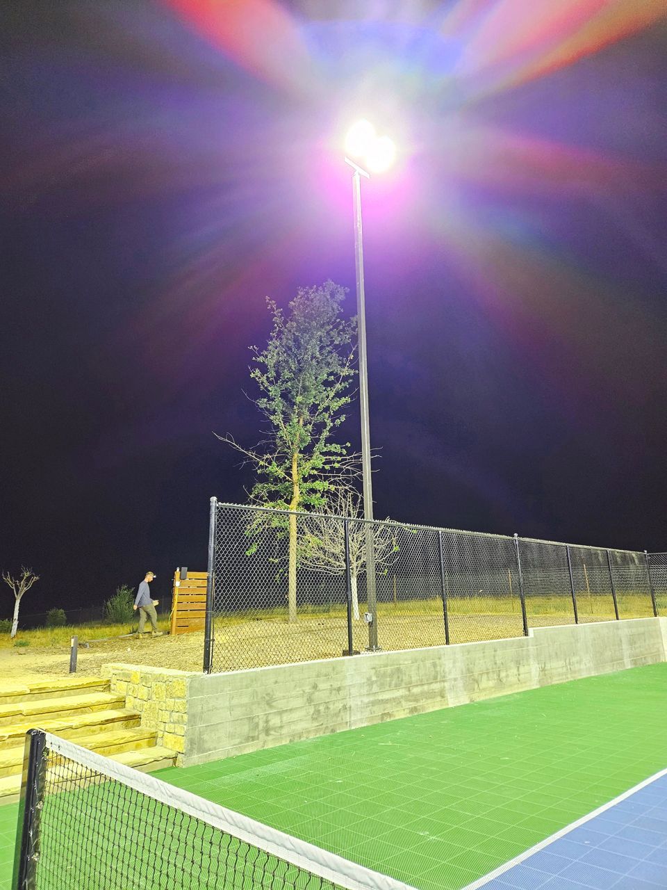 Tennis court at night with a bright overhead light, a person near the fence, and a tree.