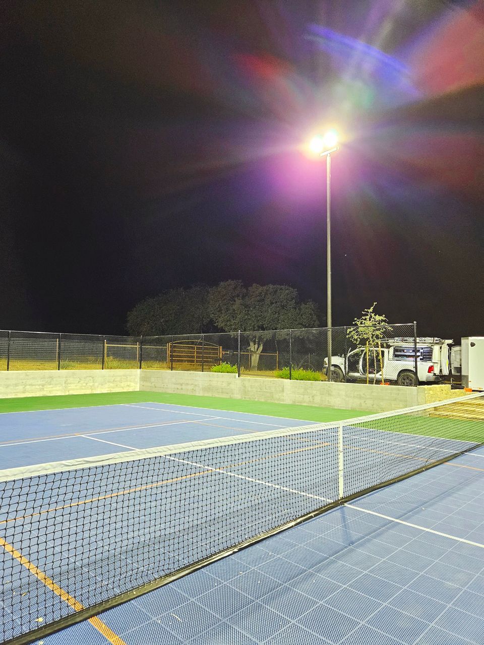 Tennis court at night, lit by bright overhead lights. Net in foreground, with fence and trees in background.