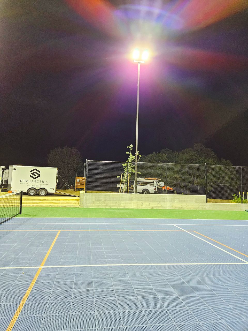 Tennis court at night, lit by bright overhead light. Trailer and equipment visible behind the court.