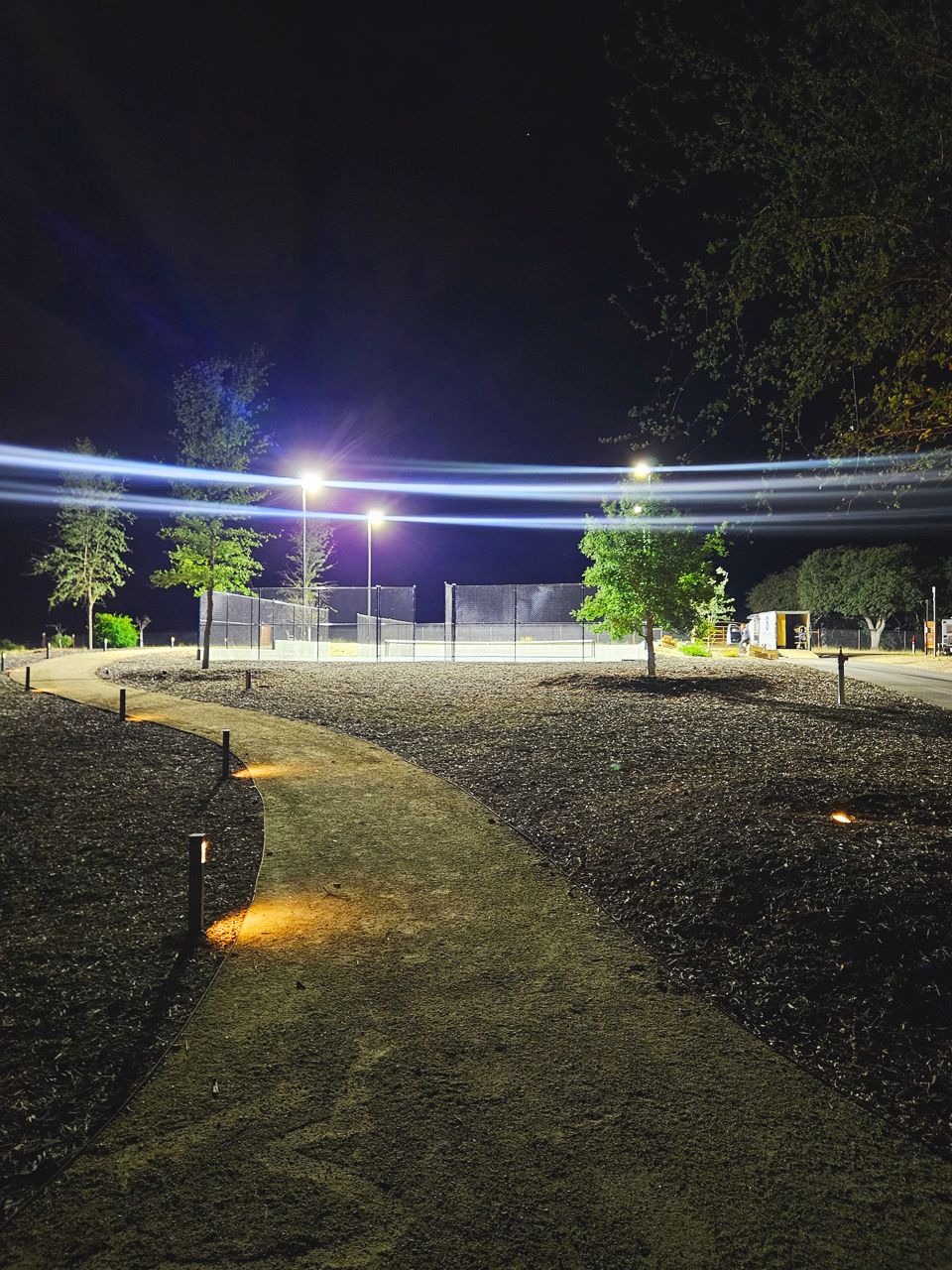 Nighttime park scene with illuminated pathway, trees, and tennis court. Long exposure captures light trails across the sky.