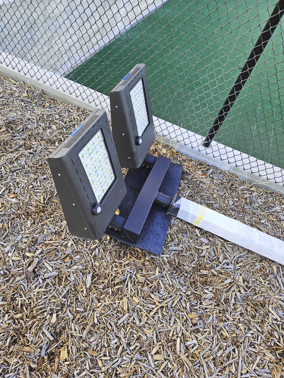 Two rectangular LED floodlights mounted on a pole, on wood chips, next to a green surface and fencing.