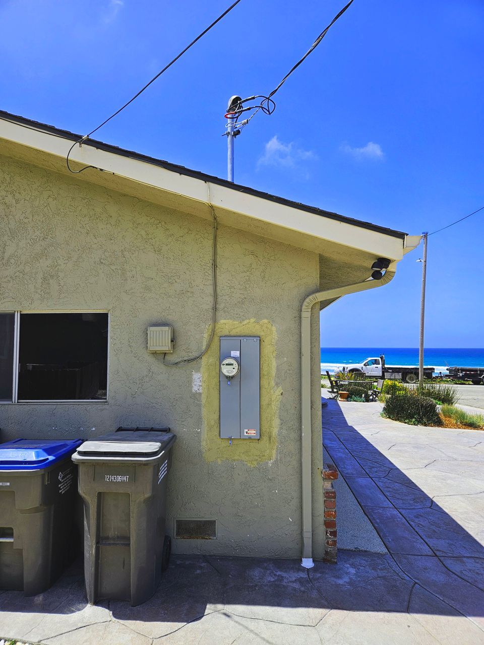 Exterior view of a stucco building with a view of the ocean. Trash cans are in front. Electric meter on the wall.
