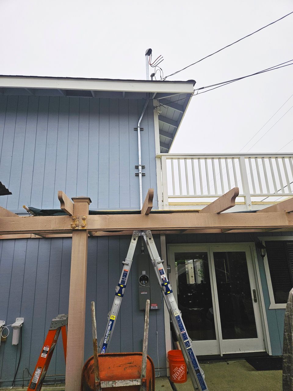 Person on a roof near power lines, painting. Ladder, pergola, and house with blue siding. Overcast sky.