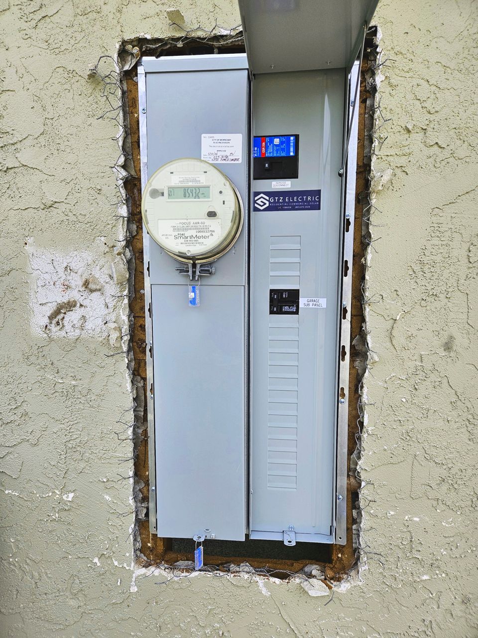 Electrical panel recessed in stucco wall, meter on the left, breaker panel on the right, door open.