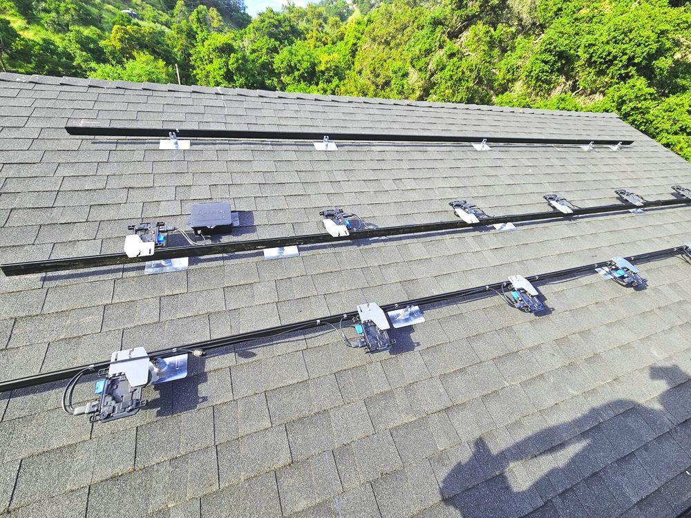 Solar panel mounting hardware installed on a dark shingled roof, trees in the background.