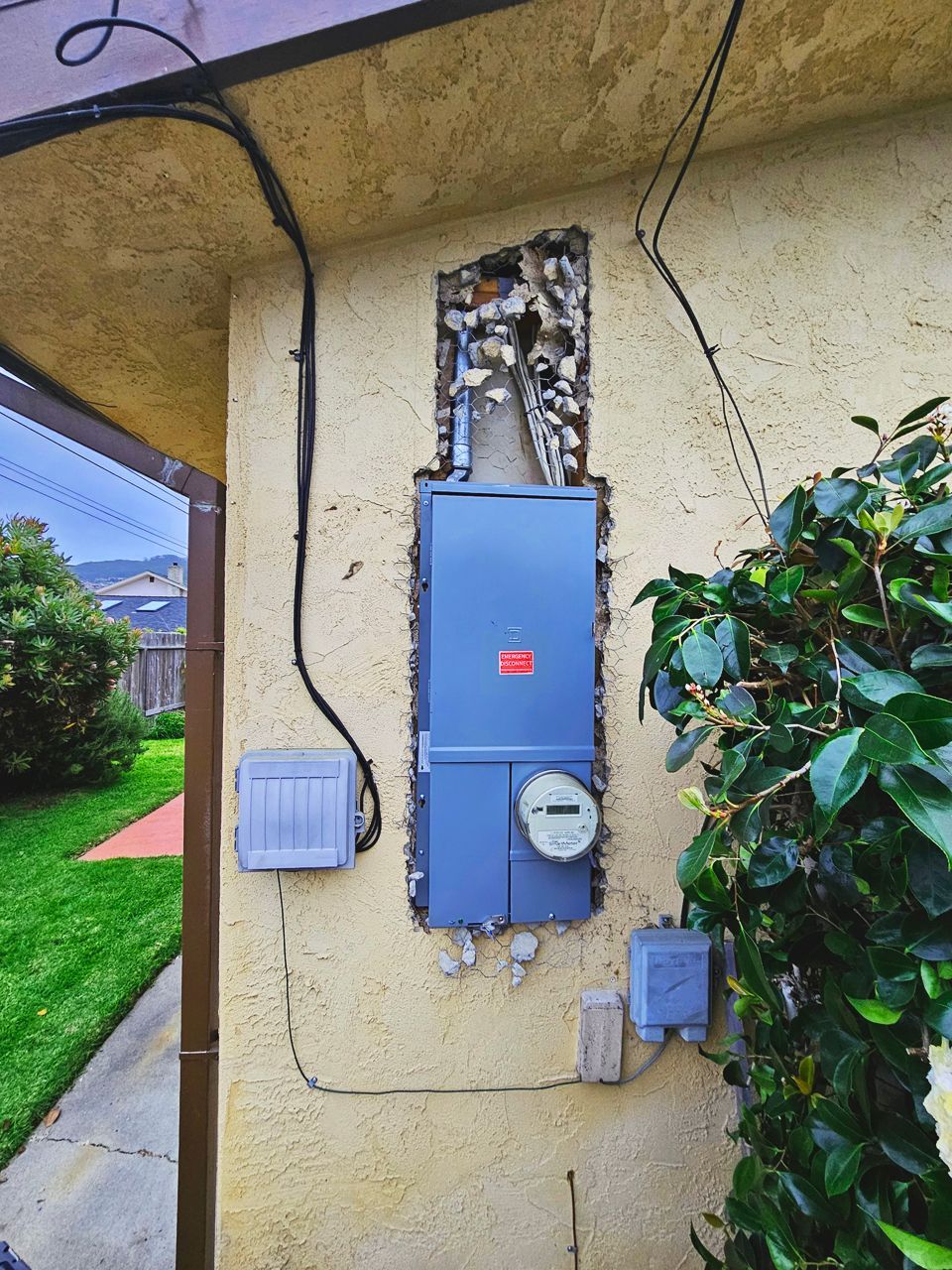 Electric meter and panel box recessed in a stucco wall, with conduit and small gray boxes, near greenery.