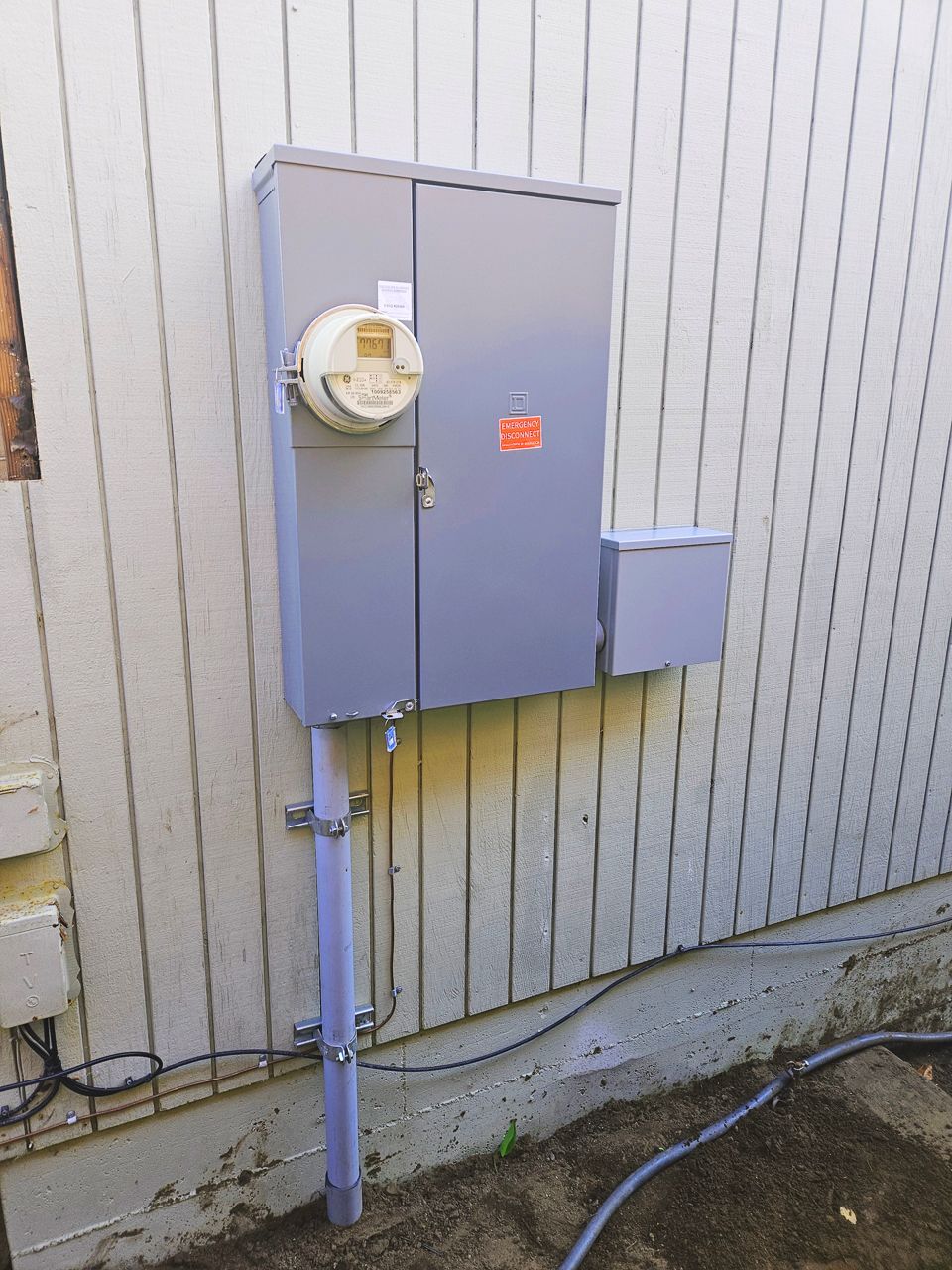 Gray electrical box with meter attached to a light-colored building. Conduit runs down to ground.