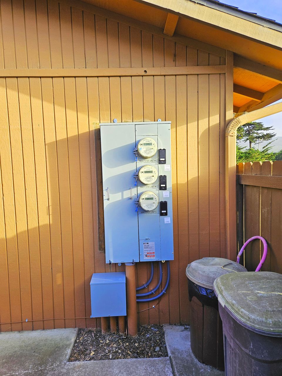 Exterior view of a gray electric meter panel on a brown wood-paneled wall, with trash cans and a concrete slab.