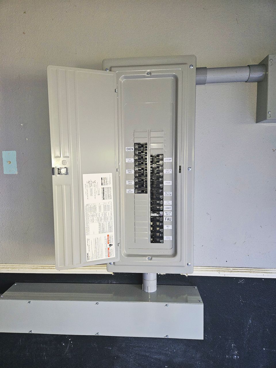 Gray electrical panel box with open door, mounted on a gray concrete base, against a gray wall.