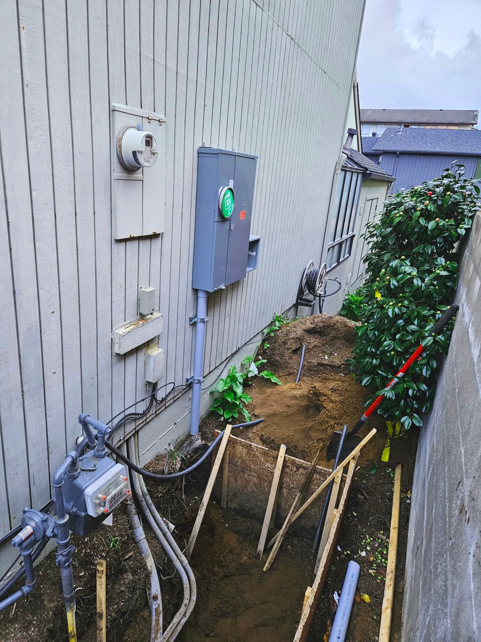 Trench dug alongside a building; electrical box, gas meter, and shovel visible. Soil, wooden stakes, overcast sky.