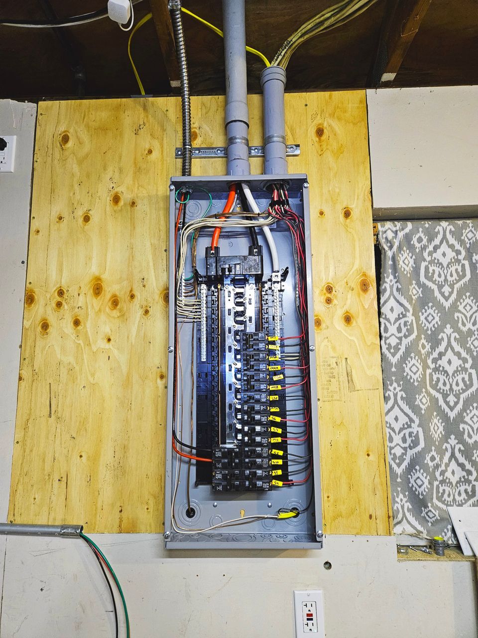 Electrical panel with wires and circuit breakers mounted on plywood. Gray conduits, white wall, and fabric curtain.