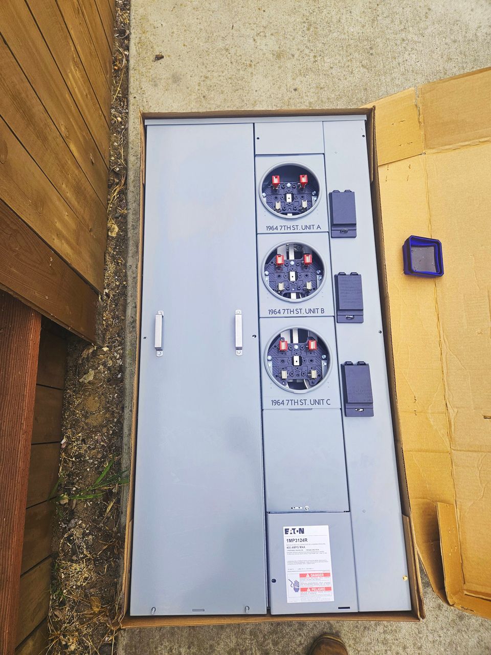 A metal electrical panel with three meter sockets, open, outdoors. Gray panel with black sockets.