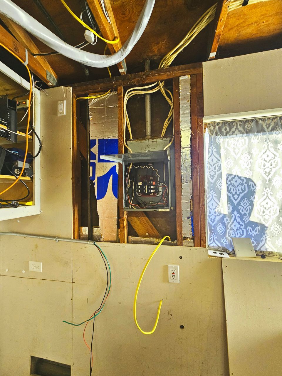 Exposed electrical panel and wiring in a wall cavity, with unfinished drywall and ceiling.