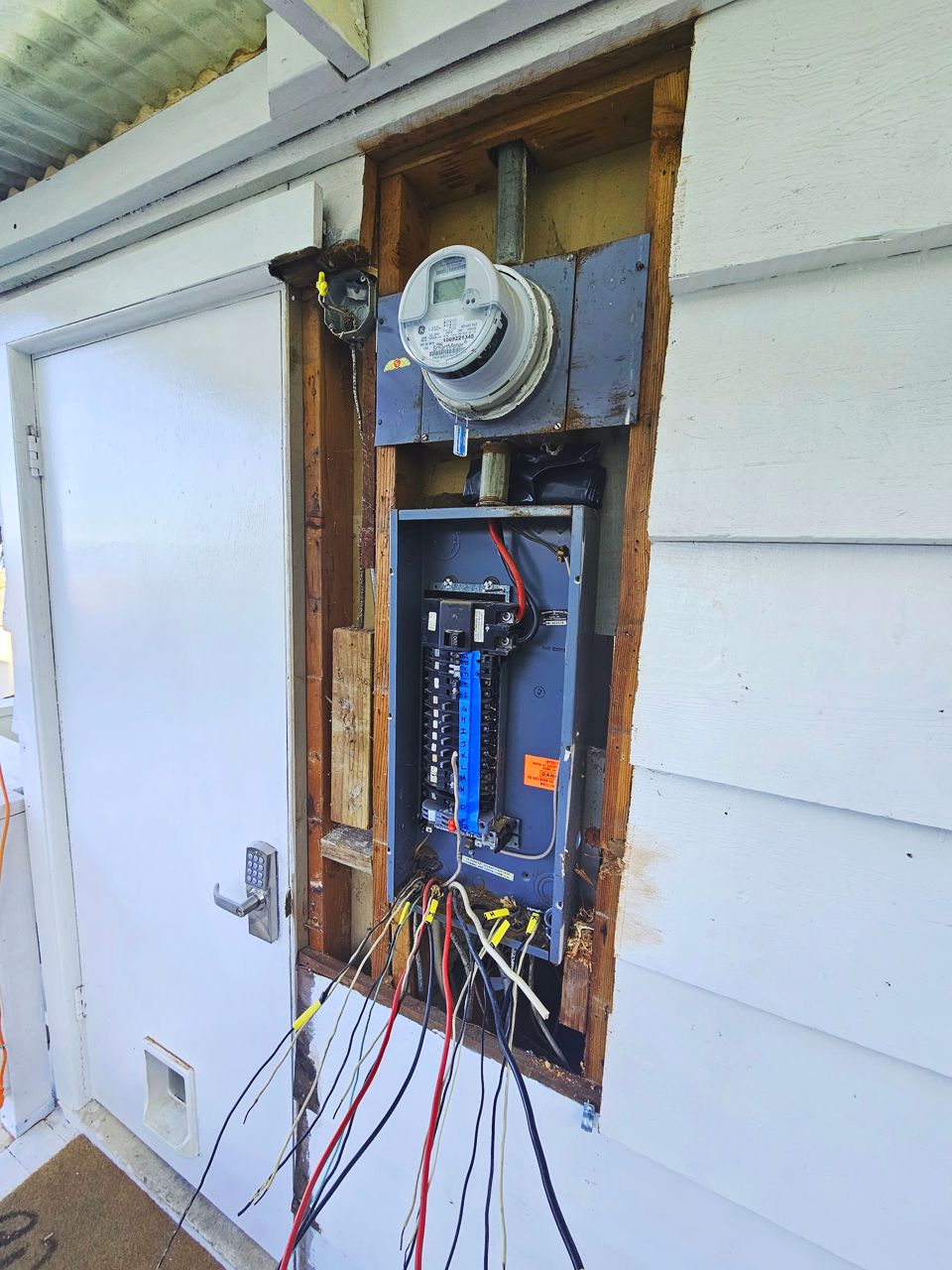 Exterior electrical panel with exposed wires and meter in a wooden frame.