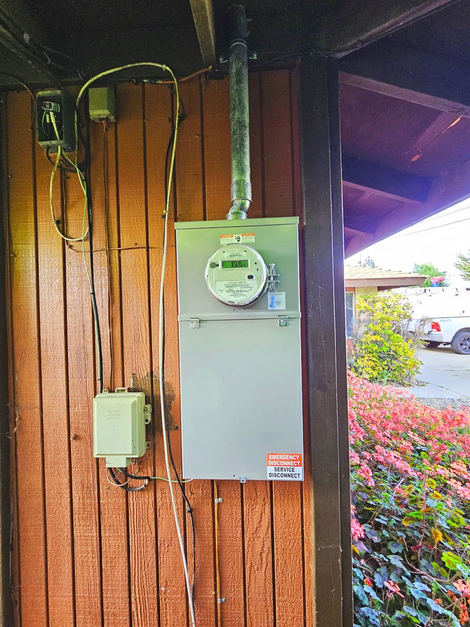 Gray electric meter box on a brown wooden wall, with wires and a green conduit.