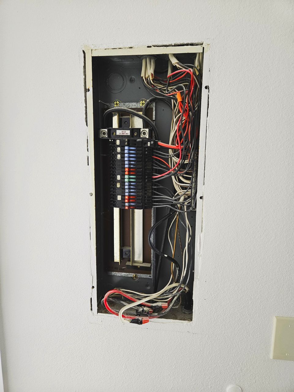 Open electrical panel recessed in white wall, filled with wires and a cylindrical component.