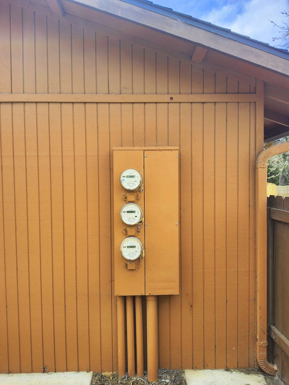 Three electric meters on a brown building's exterior, with conduit pipes at the bottom.
