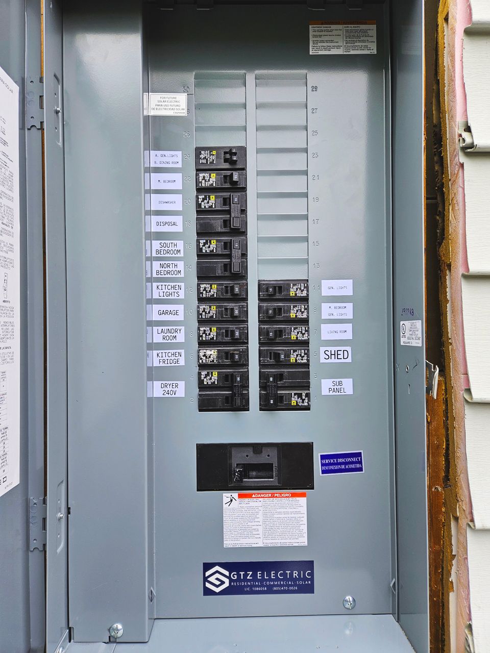 Electrical breaker box on a gray exterior wall. Several switches labeled with white stickers.