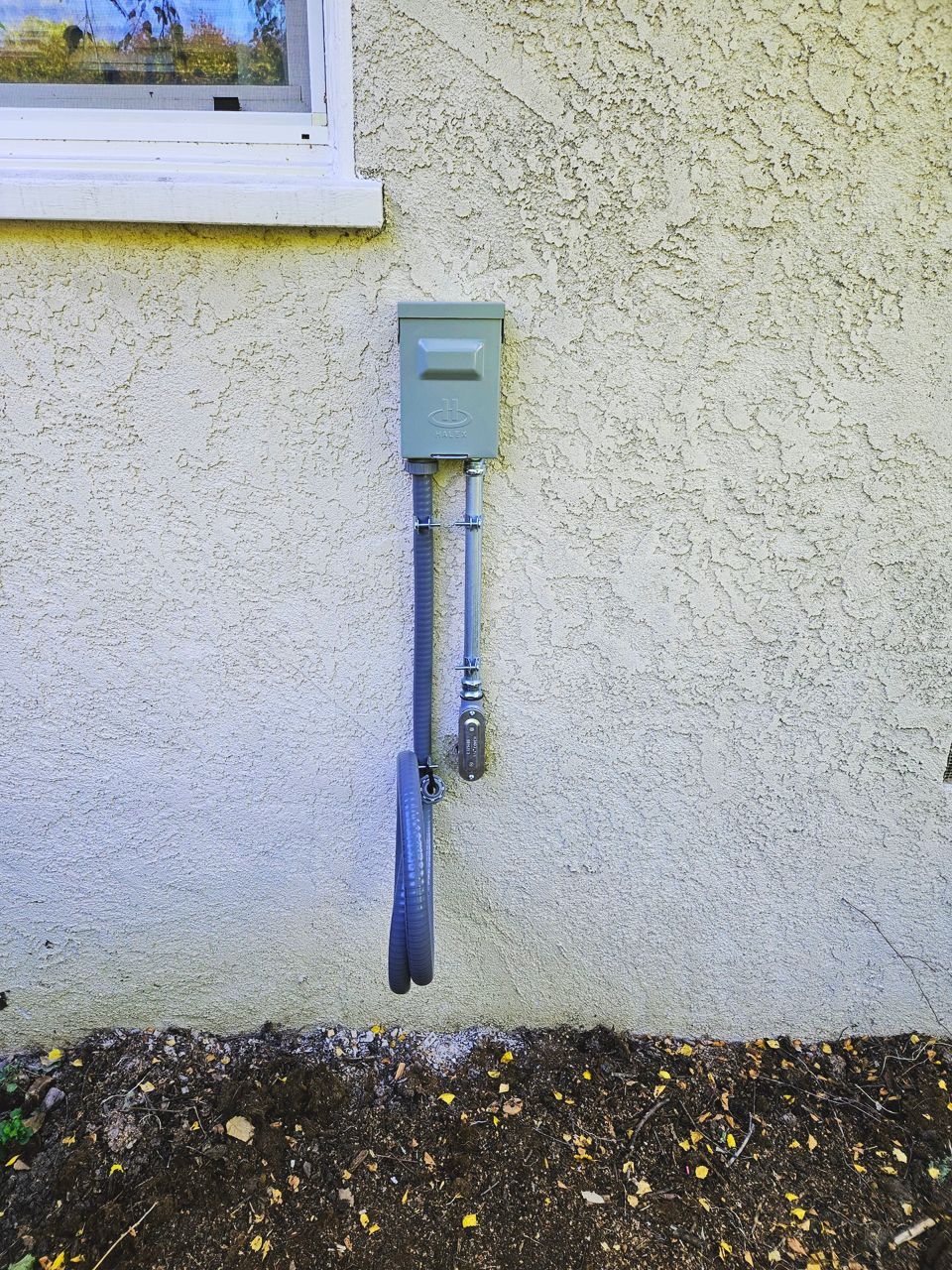 Green utility box and pipes on stucco exterior wall above a dirt patch.