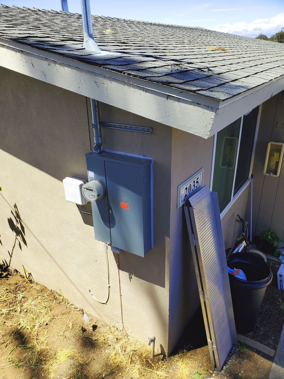 Electrical box mounted on a beige house exterior with conduit and a pipe extending above roof.