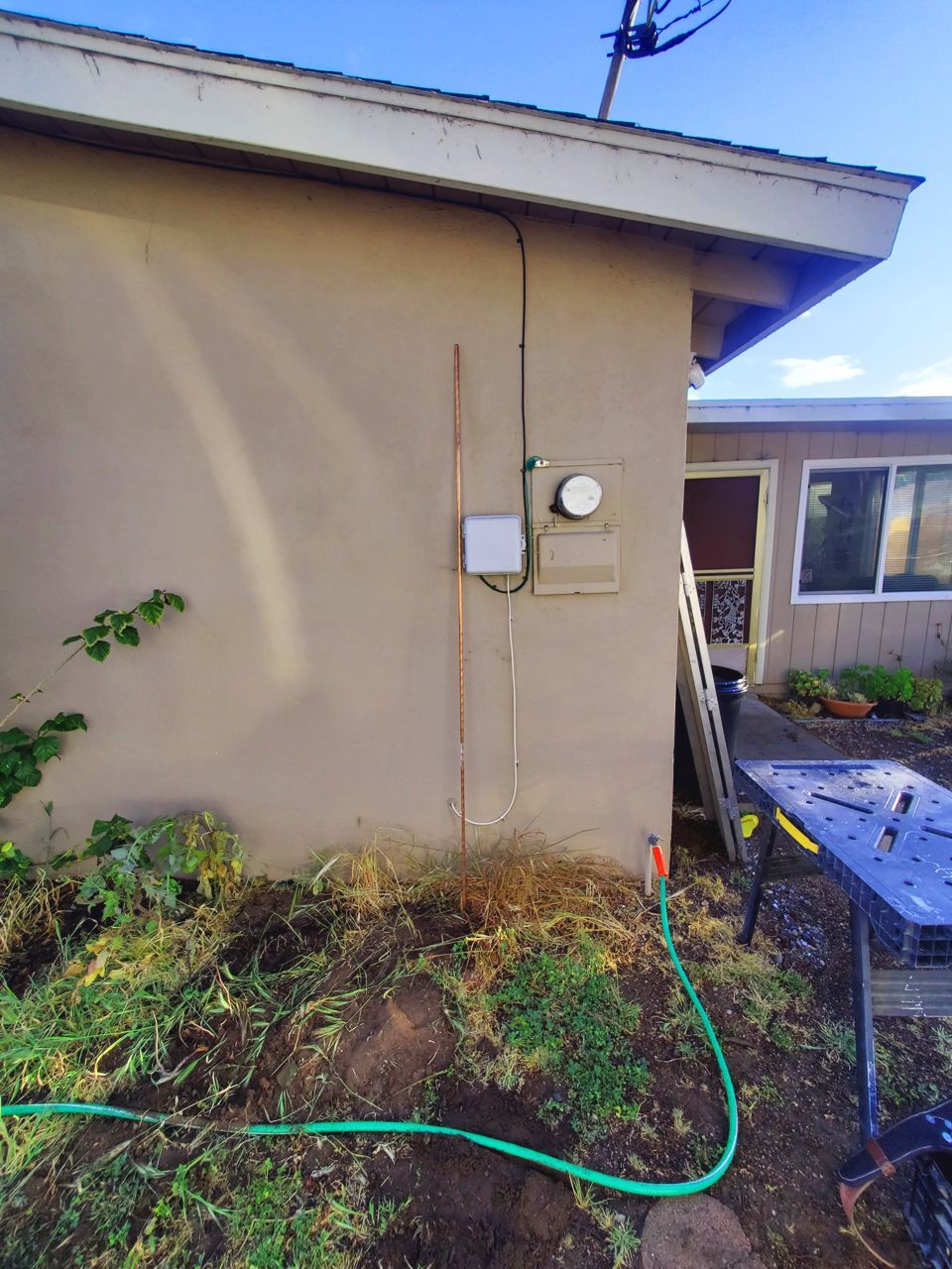 Exterior wall with electrical box, copper grounding rod, and green hose in front of overgrown vegetation.