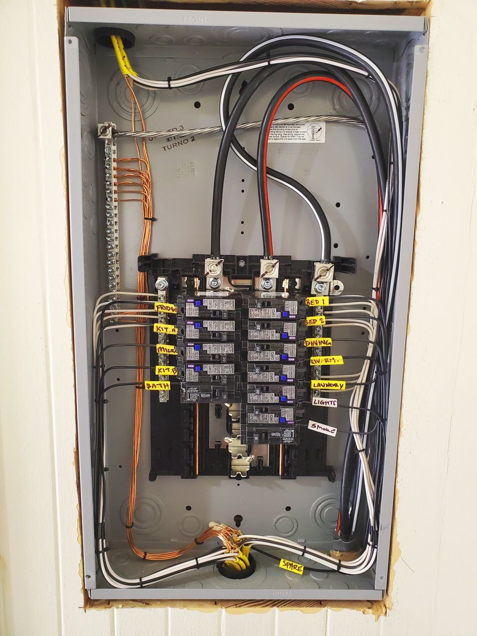 Electrical panel with wires and circuit breakers, mounted in a wall. Gray metal box, copper and black wires.