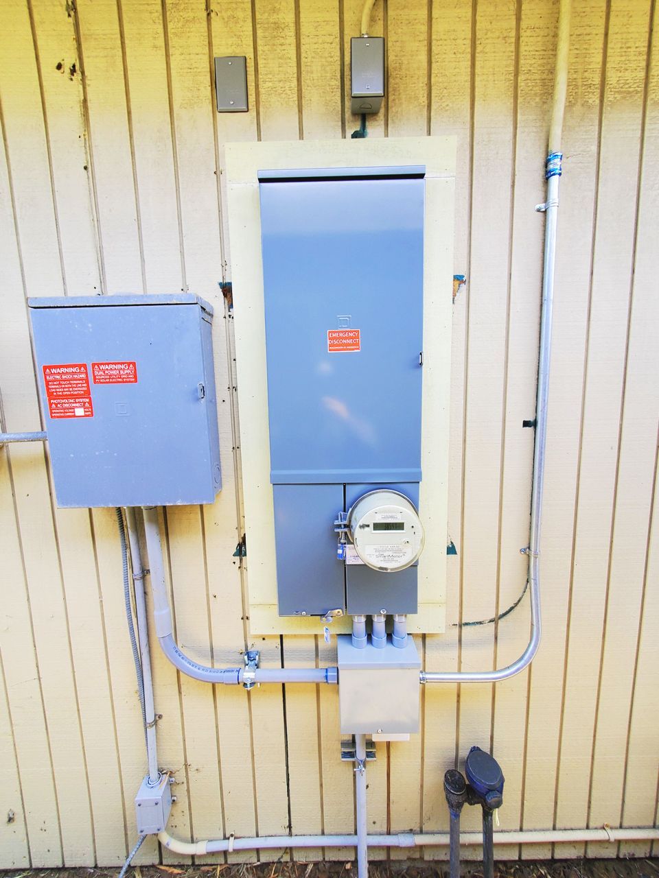 Exterior electrical panel and meter box on a yellow-sided building. Gray boxes, conduit, and meter.