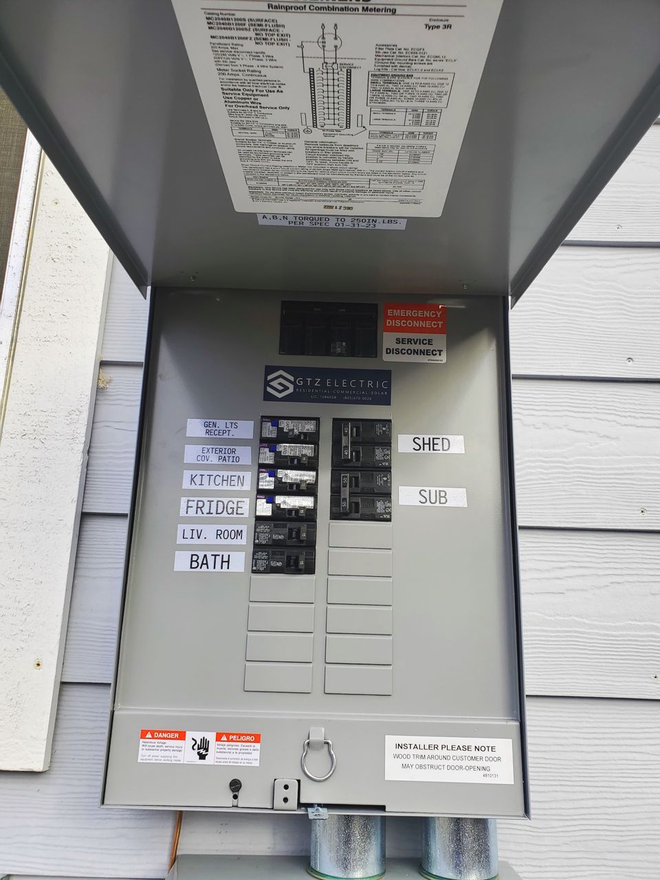 Open electrical panel on a building exterior, with labeled circuit breakers. Gray metal, white siding background.