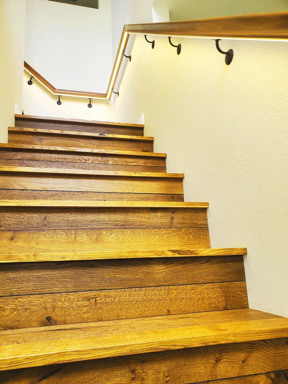 Wooden staircase with a handrail along a cream-colored wall, lit by small black light fixtures.