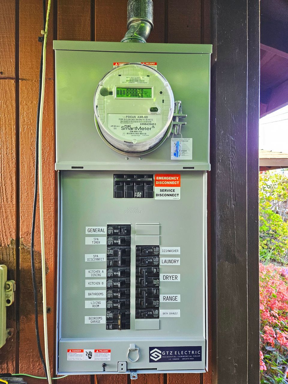 Electric meter and breaker box mounted on a wooden building exterior with circuit labels.
