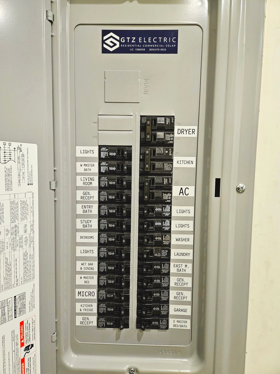 Electrical panel with labeled circuit breakers, mounted on a wall. Silver metal with gray background.