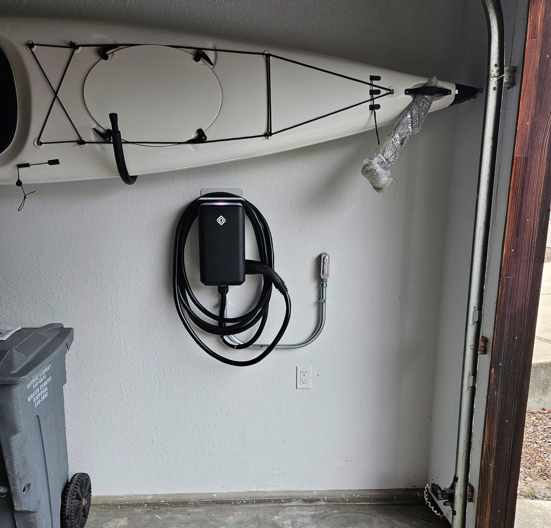 A white kayak hanging above an EV charger mounted on a garage wall, next to a trash bin.