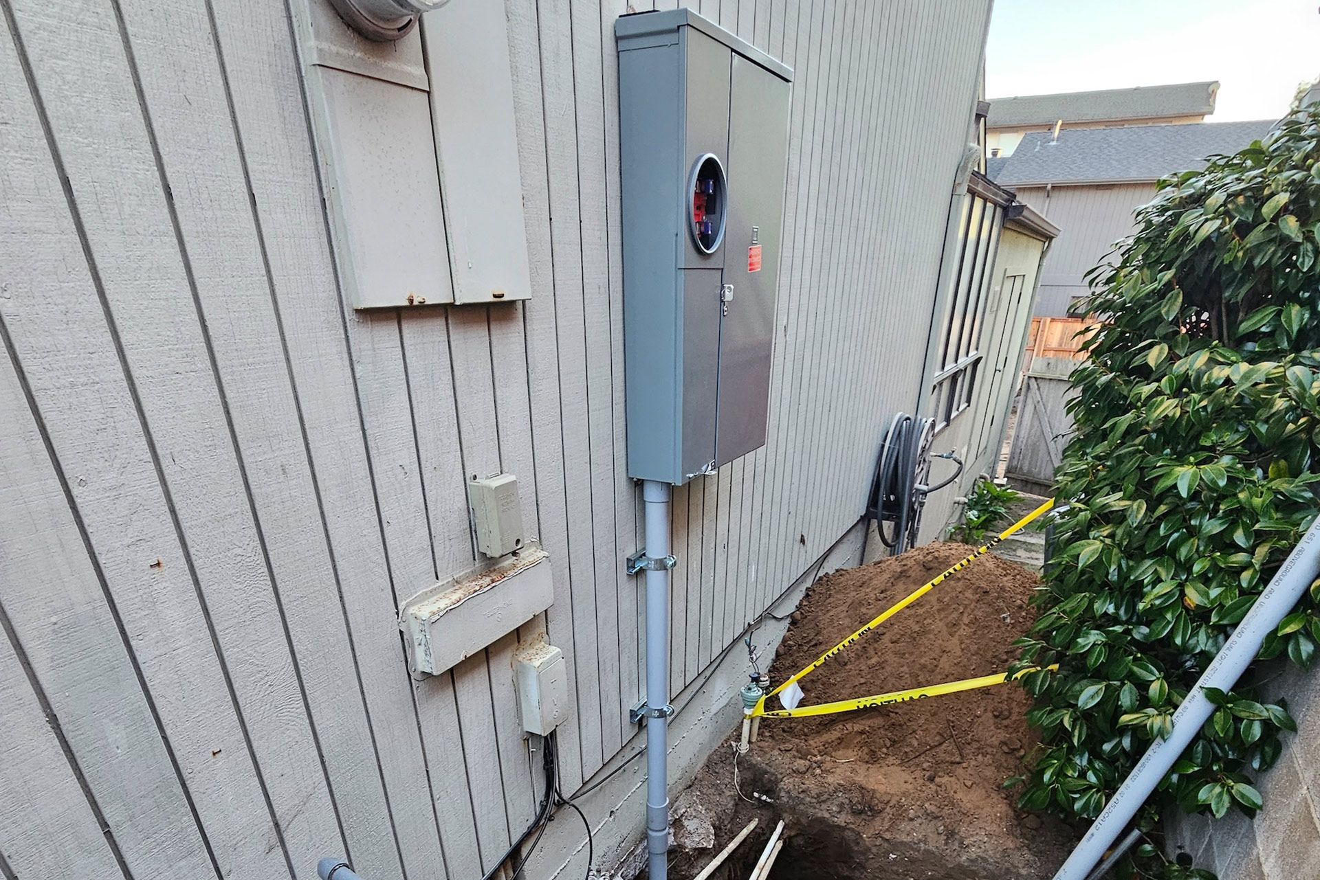 Electrical meter box mounted on a white house exterior with a small dirt pile and caution tape.