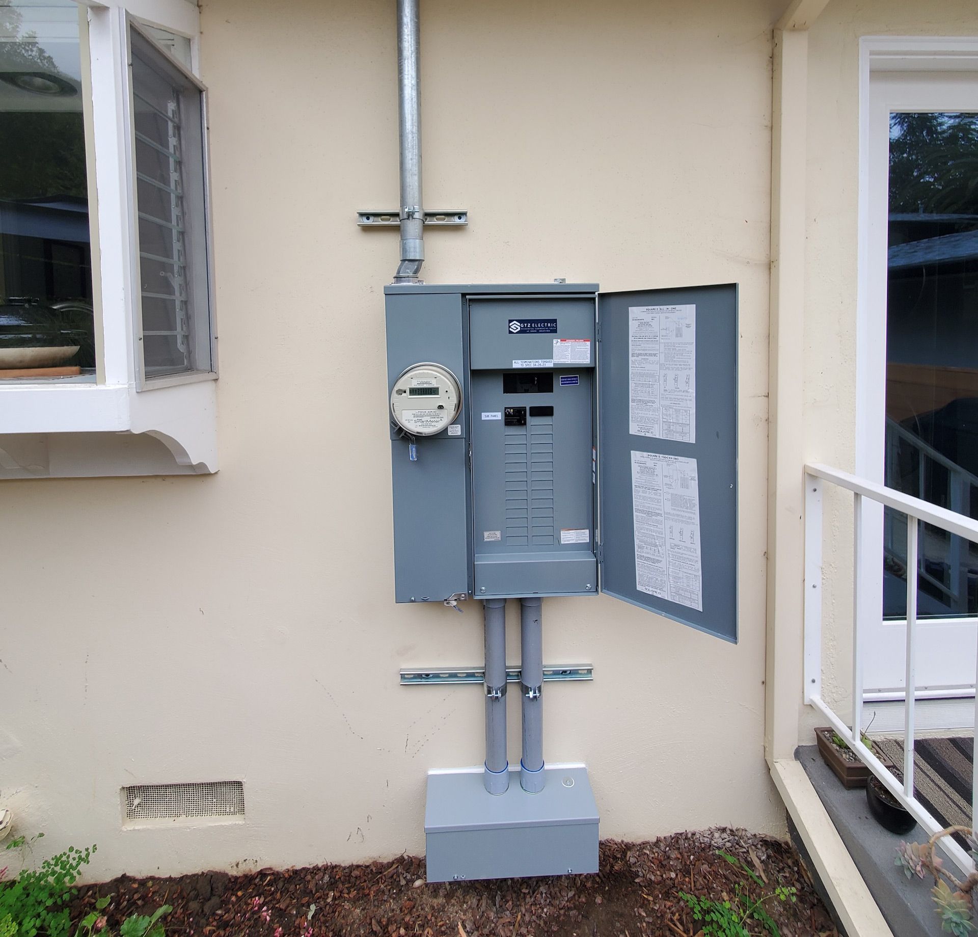 Exterior electrical panel with open door, meter, and conduits on a beige house wall.