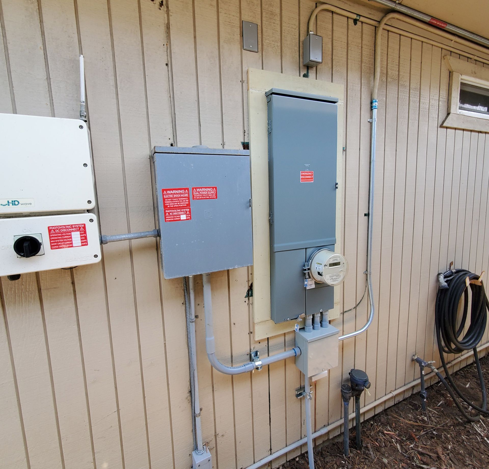 Electrical equipment on a beige building exterior: an inverter, breaker box, meter, conduit, and hose.