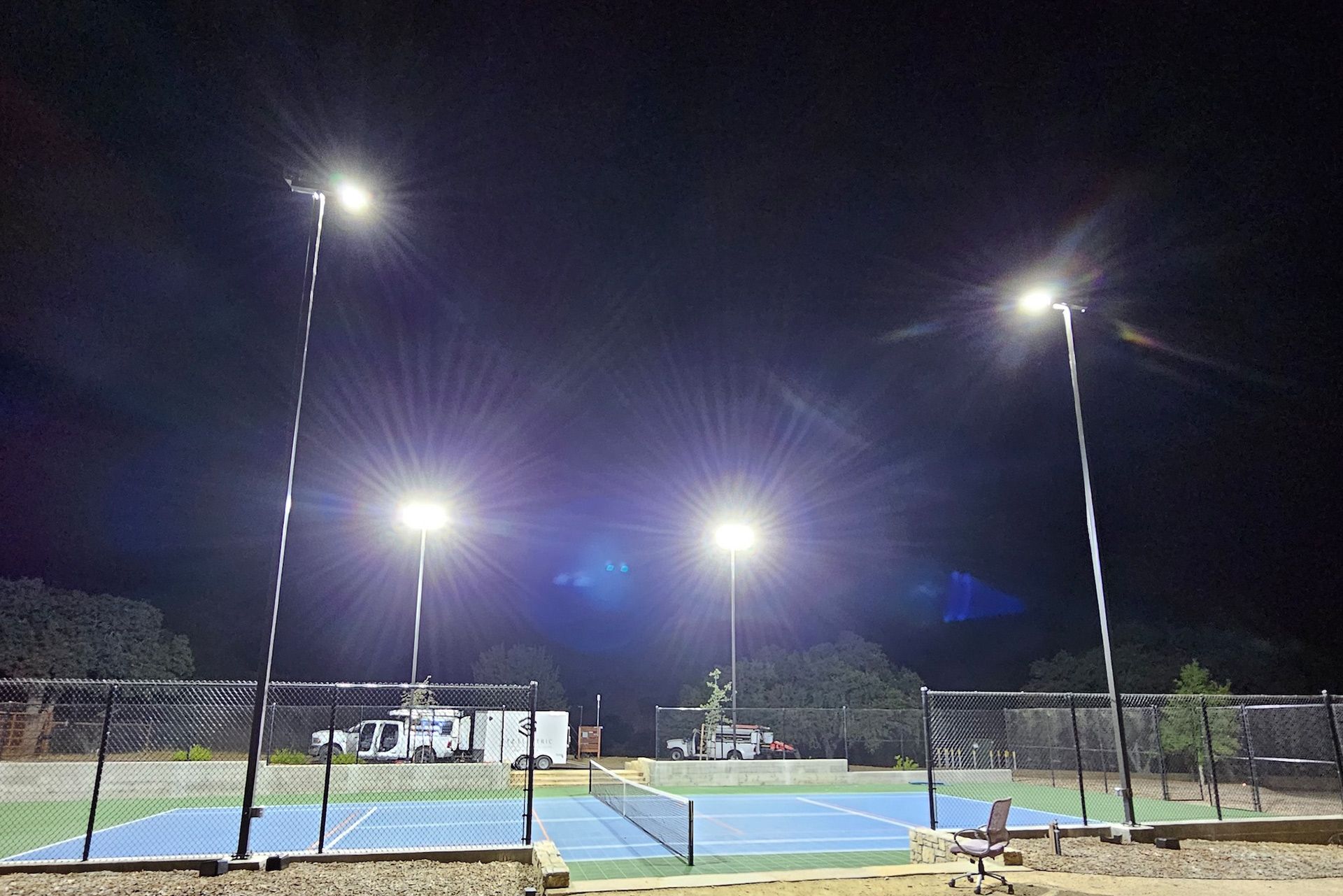 Lit tennis court at night, with two bright light poles. A blue court is enclosed by a fence.