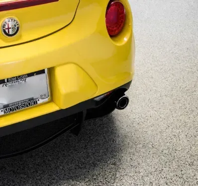 Yellow sports car's rear, showing taillight, exhaust pipe, license plate, and black mesh, set against a speckled floor.