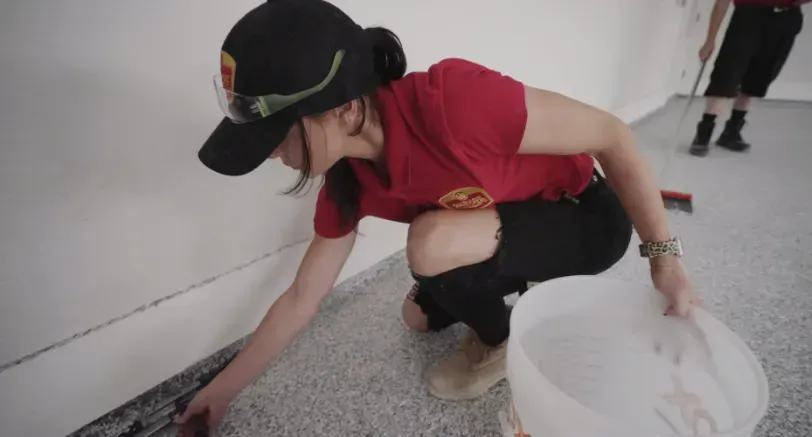 Woman in red shirt and cap applying coating to a floor, holding bucket. Indoors.