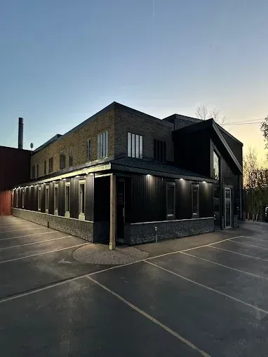 Aerial view of a long, flat-roofed building with large doors and a parking lot. Cars line the nearby road. Aerial view of a long, flat-roofed building with large doors and a parking lot. Cars line the nearby road.