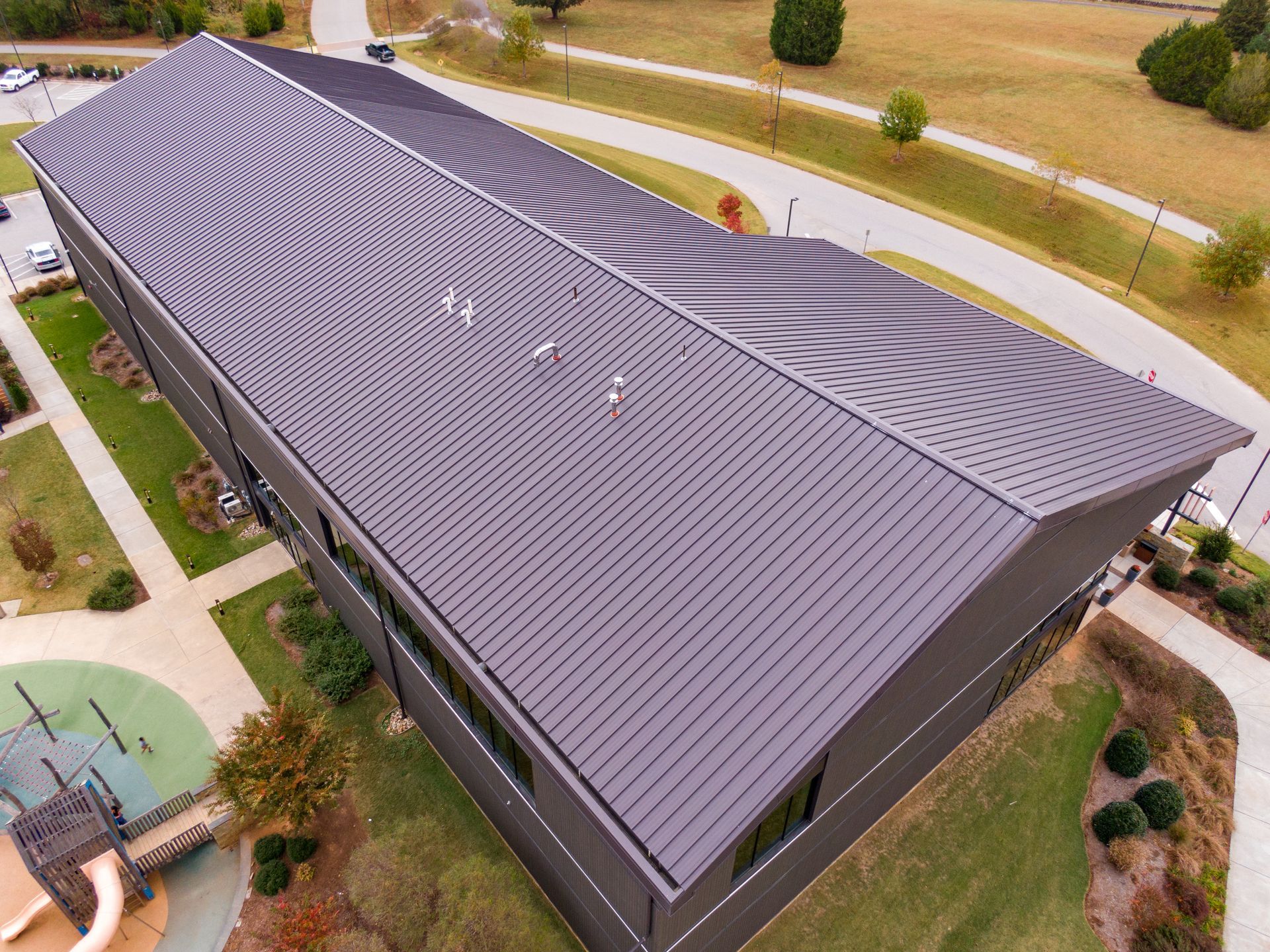 Aerial view of a dark metal-roofed building next to a grassy field and paved walkways. Aerial view of a dark metal-roofed building next to a grassy field and paved walkways.