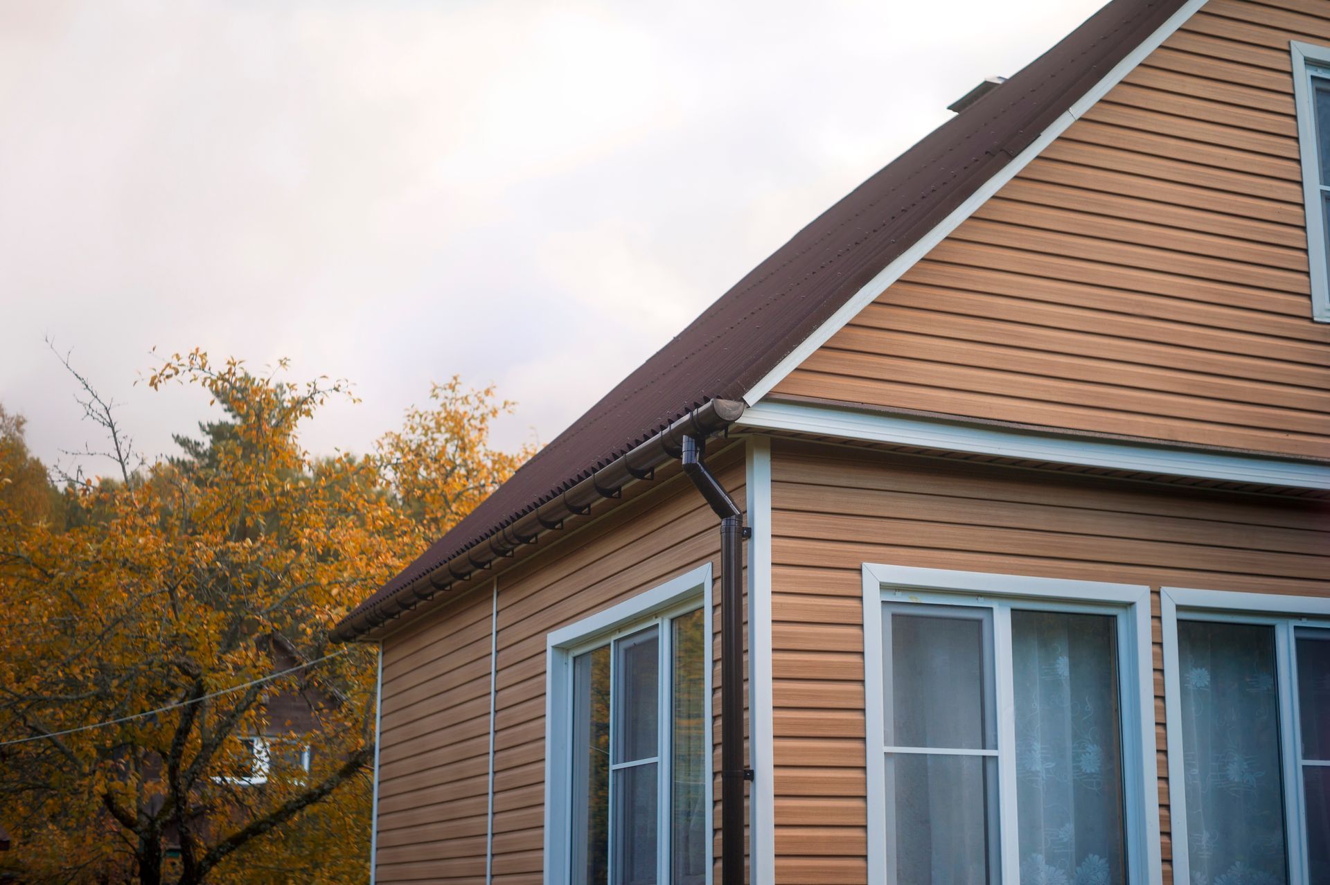 Tan-sided house with brown roof and gutters, a window, and an autumn tree in the background. Tan-sided house with brown roof and gutters, a window, and an autumn tree in the background.