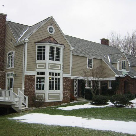 Beige and brick two-story house with bay windows, deck, and snow on the lawn.
