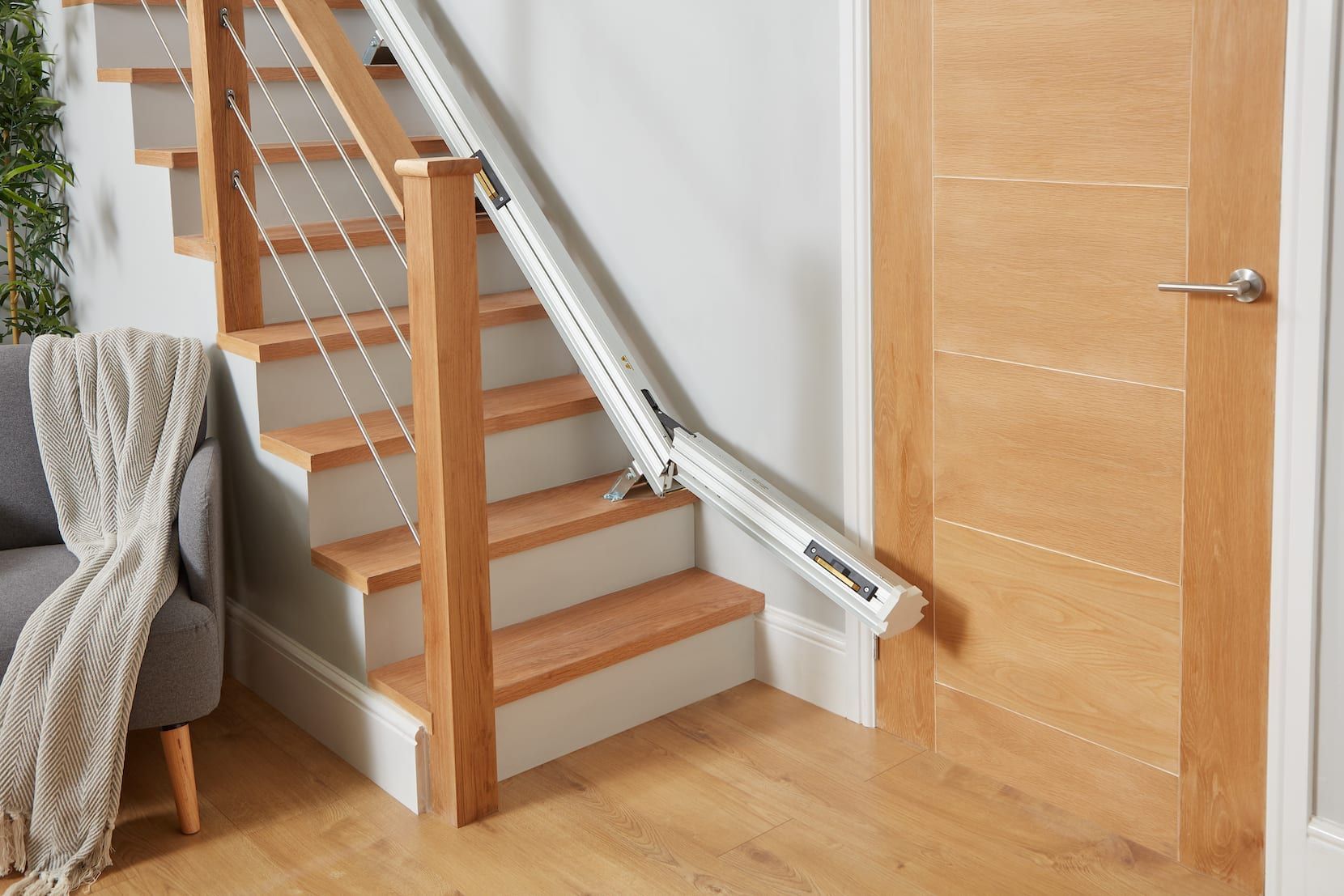 Wooden indoor staircase with a stairlift beside it, next to a closed wooden door and armchair.