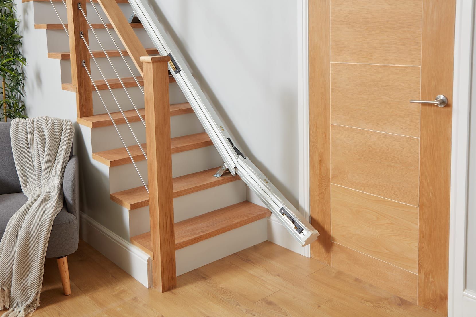 Interior staircase with a stair lift rail beside a wooden door and chair in a bright hallway