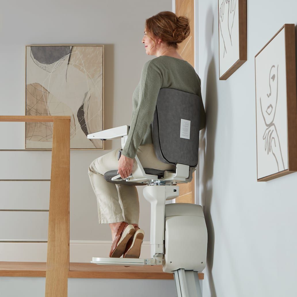 Woman seated on a stair lift in a bright home interior, shown side view moving upstairs