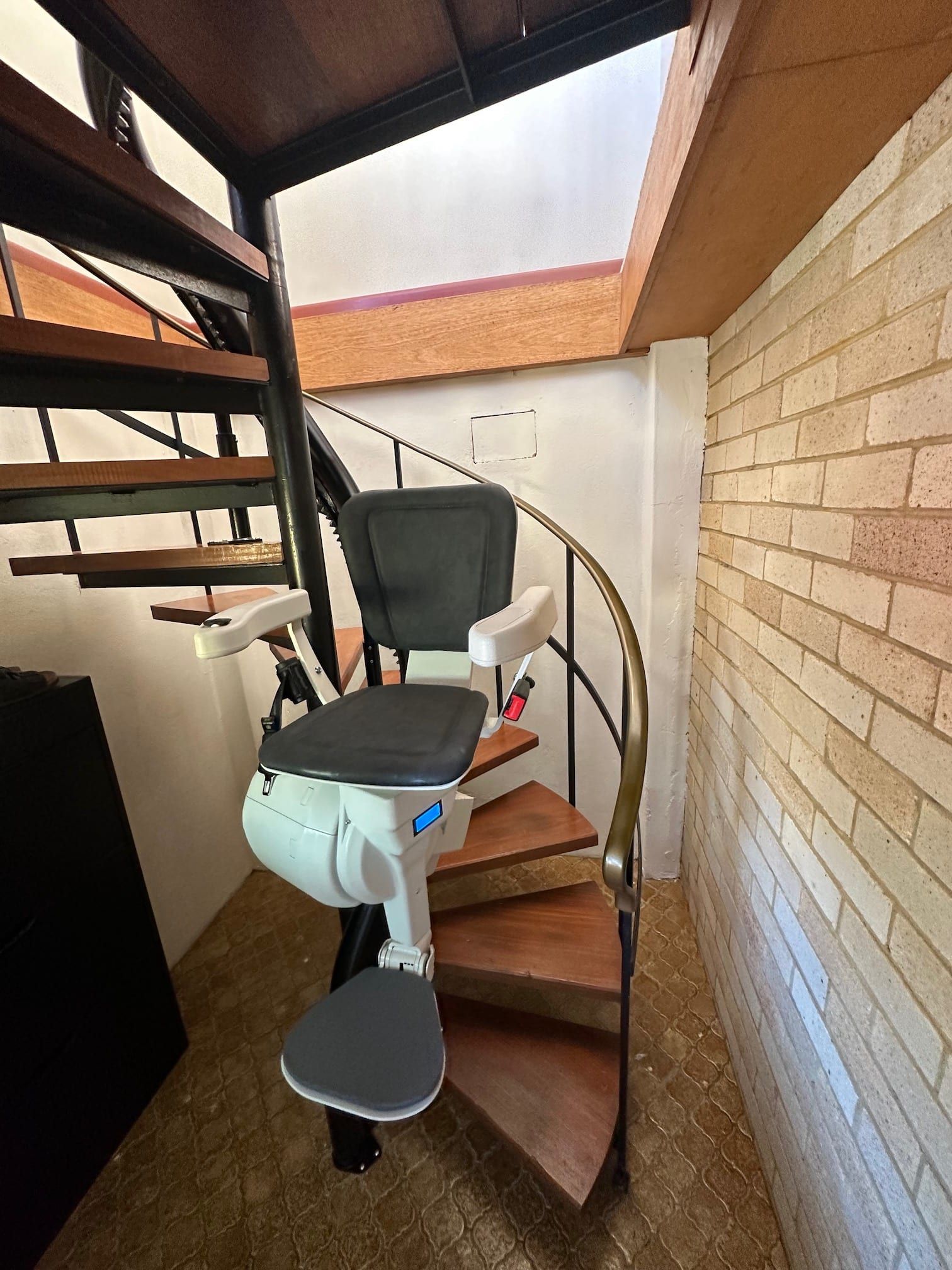Indoor stairlift on a narrow spiral staircase beside a brick wall and white doorway.