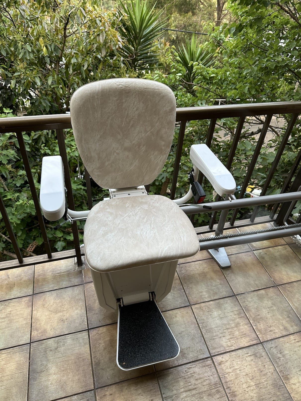 Padded outdoor transfer chair with armrests and foot platform on a tiled balcony overlooking greenery